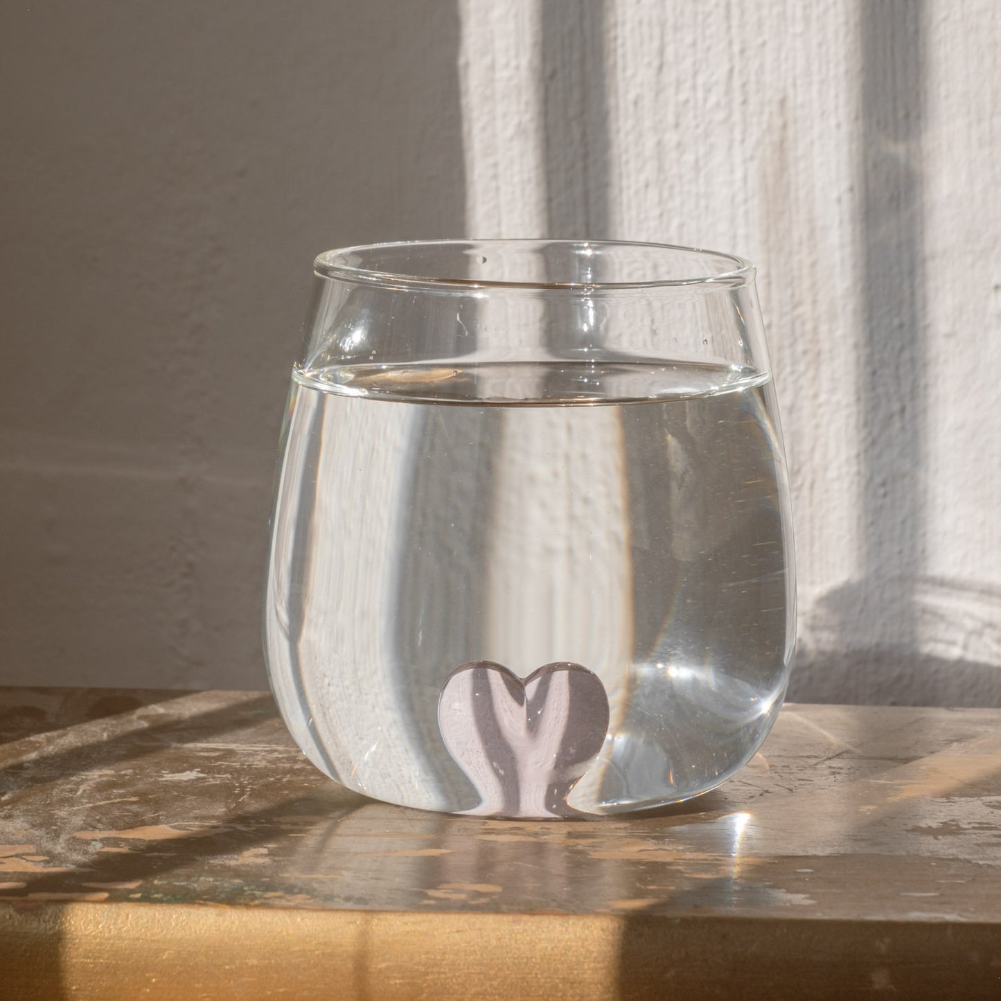 Figurglas mit rosa Herzform in der Mitte vom Glas, gefüllt mit klarem Wasser auf Holztisch vor weißer Wand.