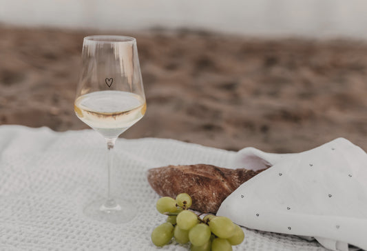 Weinglas mit Herz-Design, dazu Baguette und Trauben auf Picknickdecke am Strand.