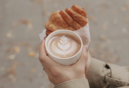 Hände halten einen Keramikbecher mit Cappuccino und Latte Art, dazu ein Franzbrötchen in Papier eingewickelt, im Hintergrund liegen herbstliche Blätter auf dem Boden.