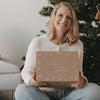 Lächelnde Frau sitzt im Schneidersitz vor einem Weihnachtsbaum und hält die limitierte Winter-Überraschungsbox 2026 von Eulenschnitt auf ihrem Schoß.
