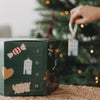 Eulenschnitt Adventskalender vor dem Weihnachtsbaum mit Baumanhänger in der Hand