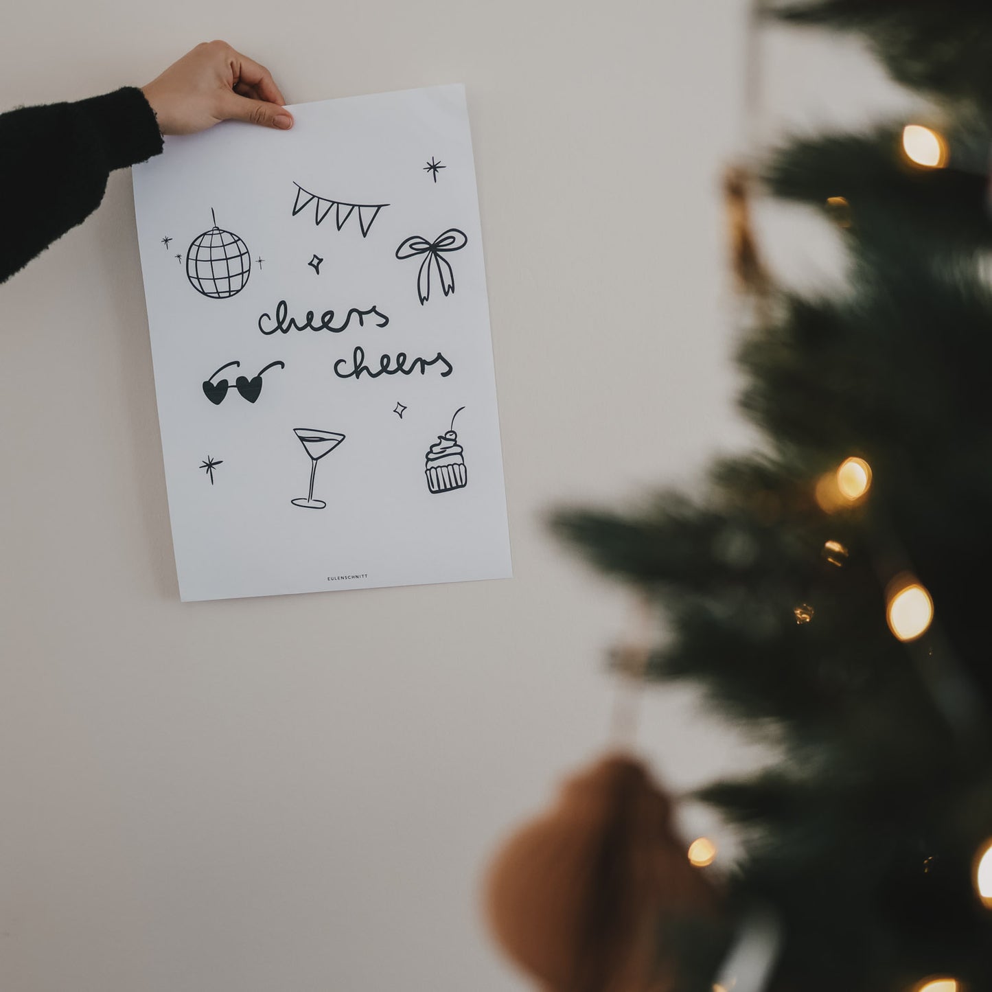 Hand hält neben Weihnachtsbaum das Winter cheers Poster in hoch. Stilvolles Design-Statement für jedes Zuhause. Eulenschnitt Poster zum Download.