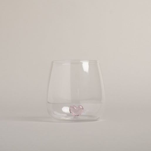 Klares Trinkglas mit rosa Herzfigur am Boden, modernes Design auf hellem Hintergrund fotografiert.