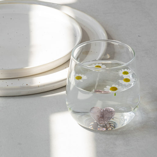 Trinkglas Herzfigur rosa mit Wasser und fünf kleinen Gänseblümchen, auf grauem Untergrund neben zwei Steingut Tellern.