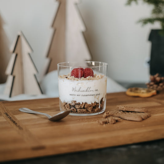 Trinkglas mit Weihnachtsgravur ‚Weihnachten ist, wenn wir zusammen sind‘ gefüllt mit Dessert, dekorativ angerichtet auf Holzbrett mit Spekulatius und Beeren