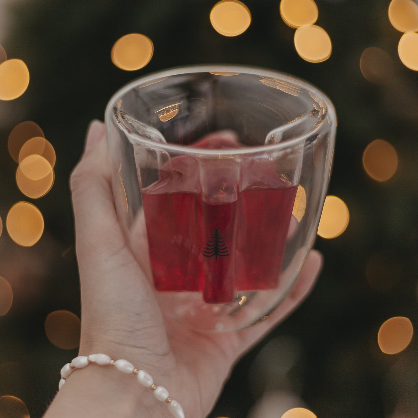Hand hält doppelwandiges Tannenglas mit rotem Getränk vor weihnachtlicher Lichterkulisse.