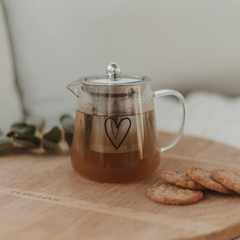 Teekanne aus Glas mit schwarzem Herzmotiv, gefüllt mit Tee, auf Holztablett mit Keksen und Eukalyptuszweig