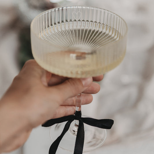 Hand hält elegantes Champagnerglas mit Herzgravur und schwarzer Schleife am Stiel.