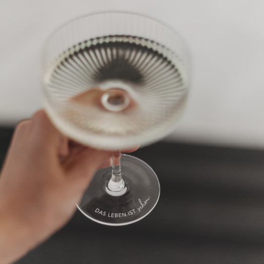Hand hält Champagnerglas mit Gravur „Das Leben ist schön“ – Blick durch das Glas von oben.
