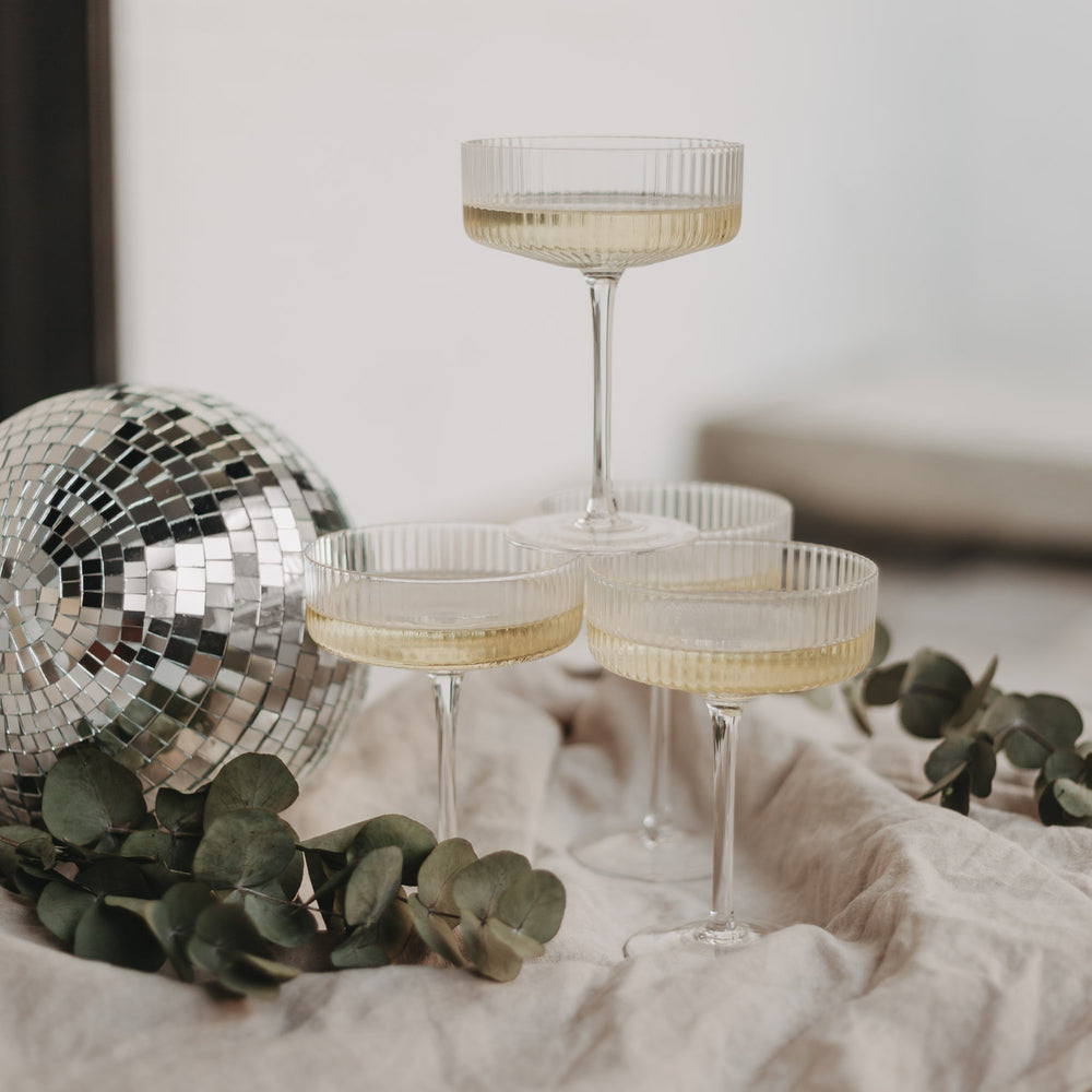Vier Champagnergläser auf Tuchdecke neben Eukalyptus und Spiegelkugel – festliche Deko.