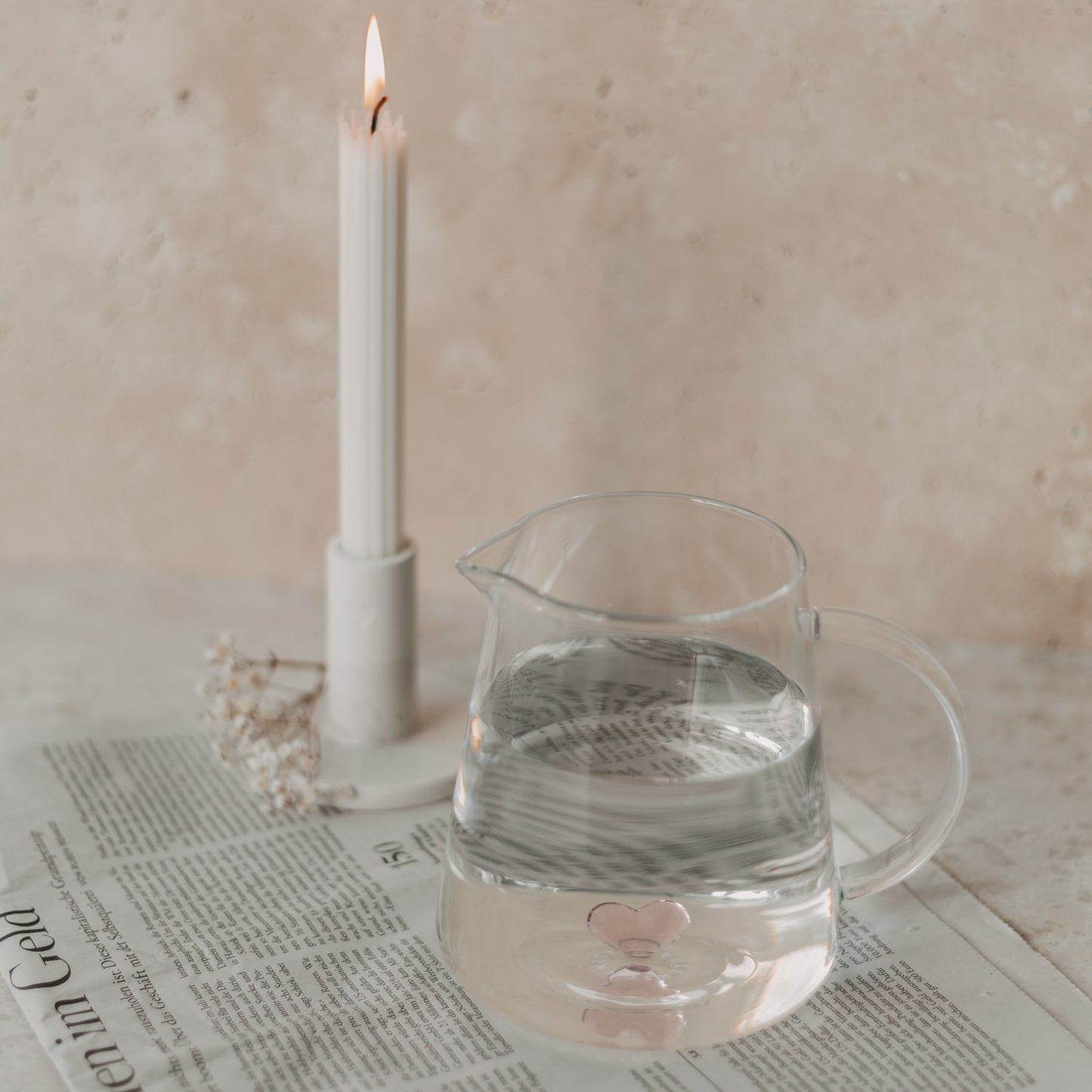 Glaskaraffe mit rosa Herz neben Kerze auf Zeitung – stimmungsvolle, liebevolle Tischdeko.