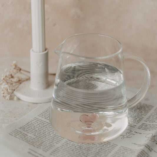 Karaffe mit Wasser und rosa Herz auf Zeitung, neben Kerzenständer mit Herzdetail.