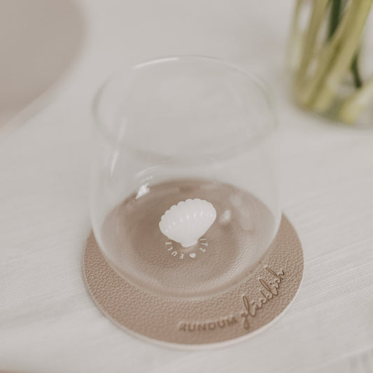 Trinkglas mit Muschelmotiv am Boden auf beigem Untersetzer mit Prägung