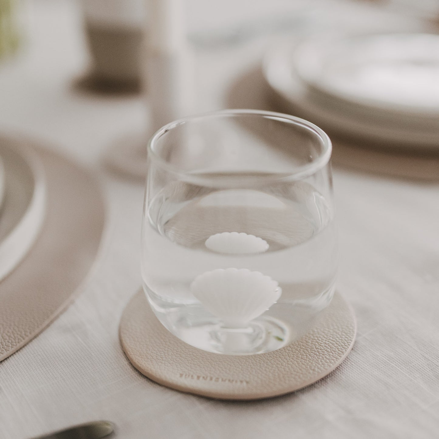 Glas mit Wasser und Muschelmotiv am Boden auf Untersetzer im natürlichen Wohnambiente