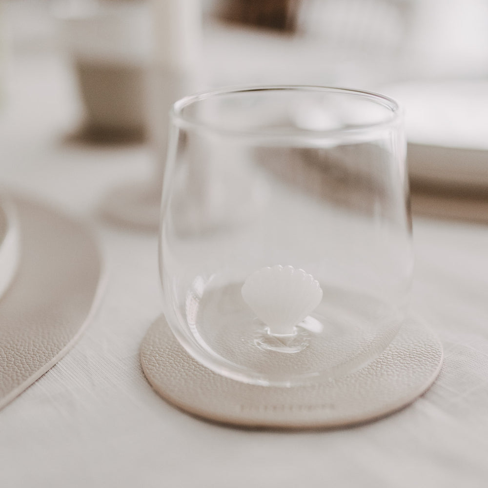 Modernes Trinkglas mit Muschelmotiv auf beigem Untersetzer in stilvoller Tischszene