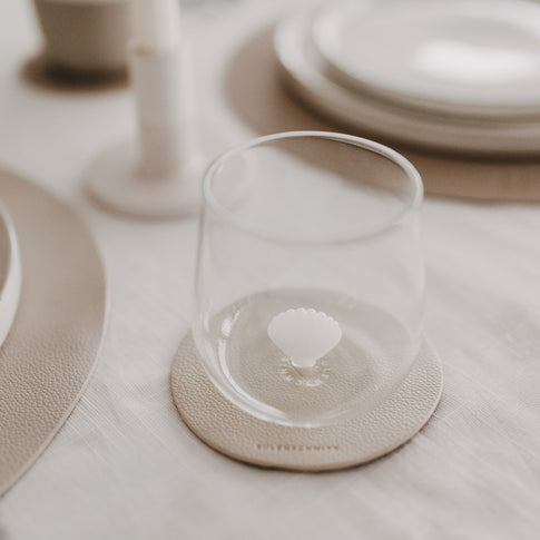 Minimalistisches Glas mit Muschelmotiv am Boden auf Untersetzer im natürlichen Setting