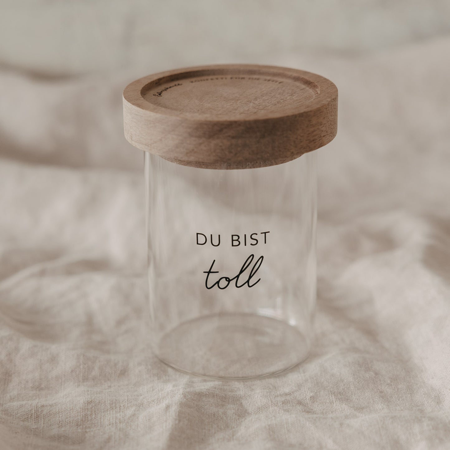Vorratsglas mit Holzdeckel und Aufdruck „Du bist toll“ auf hellem Stoff