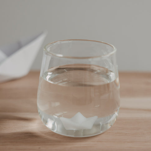 Trinkglas mit Papierboot Motiv gefüllt mit Wasser, schlichtes Glas auf Holzoberfläche