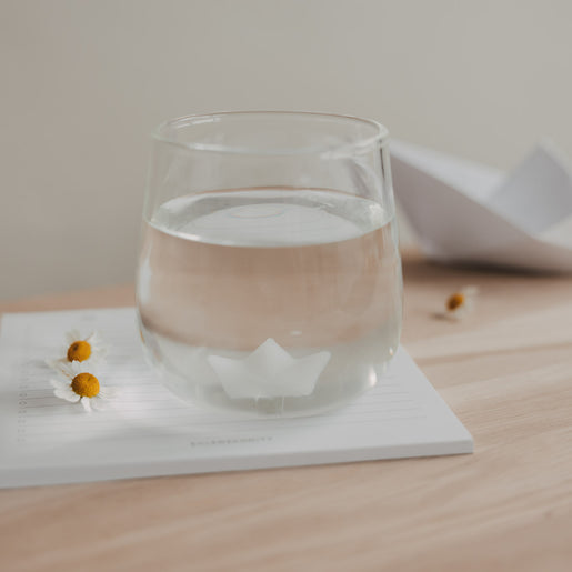 Glas mit Papierboot Motiv und Wasser auf Notizblock, dekoratives Trinkglas mit Blüten