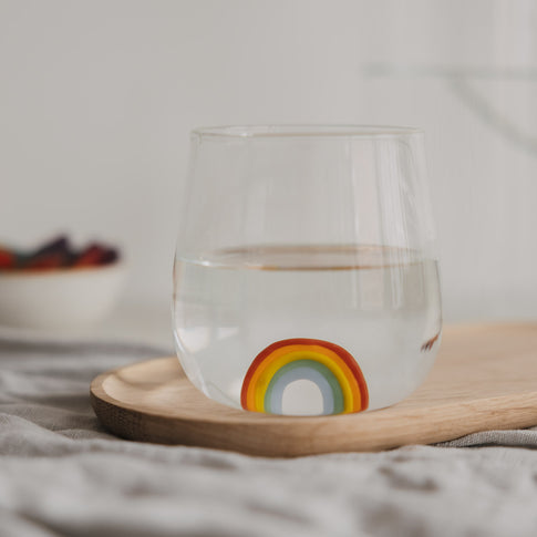 Trinkglas mit Regenbogen Motiv gefüllt mit Wasser, dekorativ auf Holzbrett arrangiert