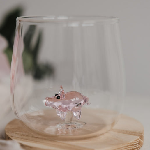 Minimalistisches Trinkglas mit rosa Schwein Figur im Boden, dekoratives Glas auf Holz