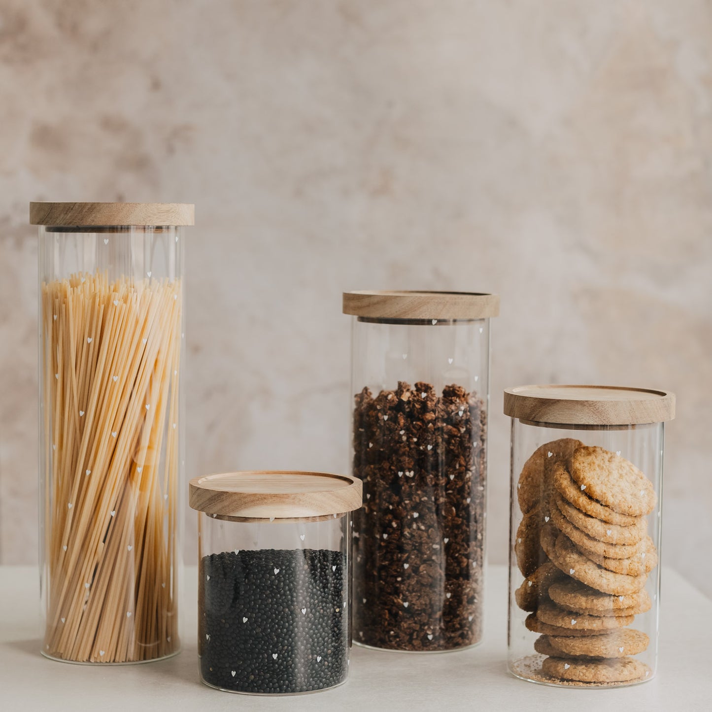 Vier Vorratsgläser mit weißen Herzen und Holzdeckeln, gefüllt mit Spaghetti, Linsen, Müsli und Keksen
