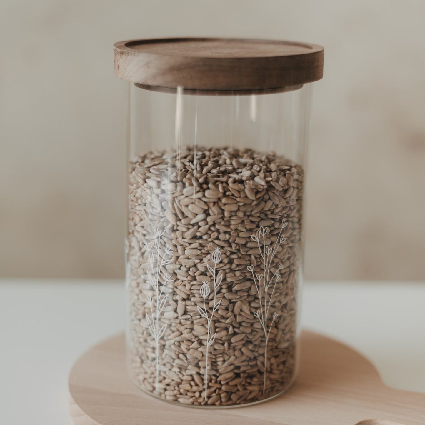 Glas-Vorratsbehälter mit feinem weißem Blumenmotiv und dunklem Holzdeckel, gefüllt mit Sonnenblumenkernen