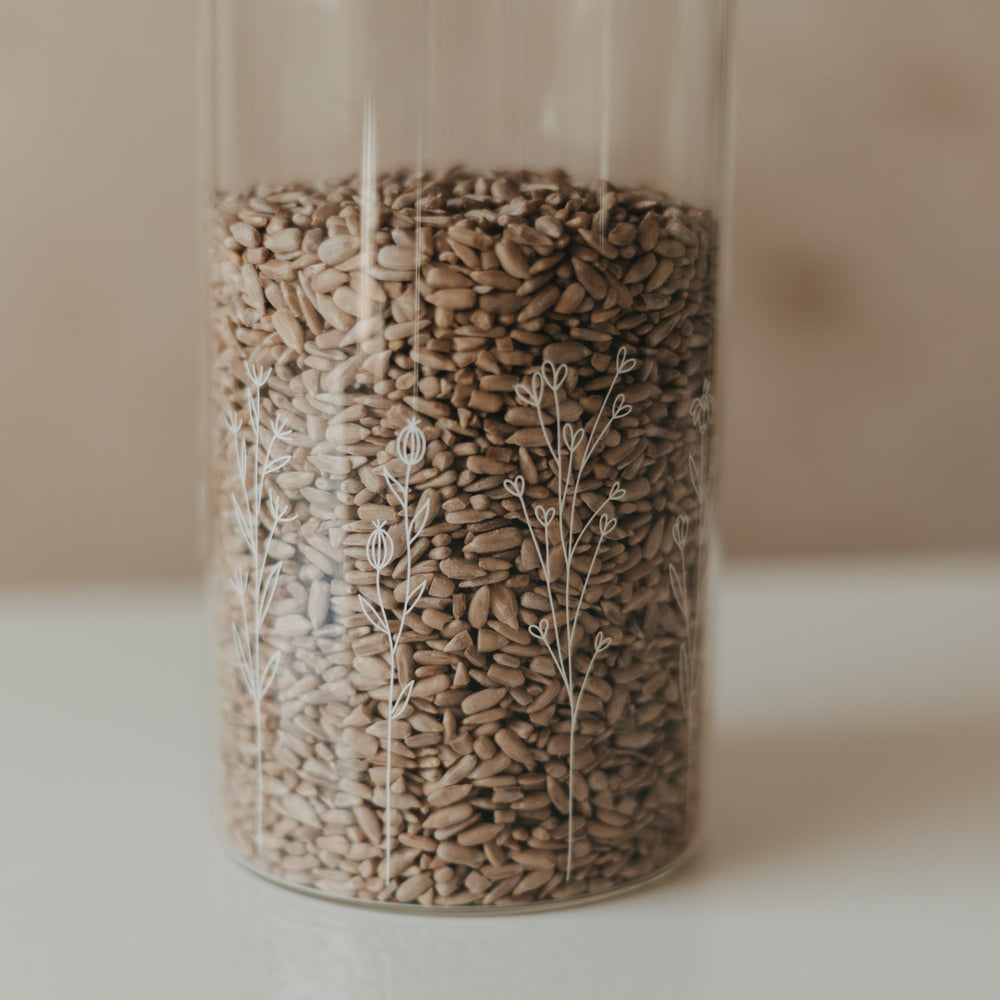 Detailansicht eines Glas-Vorratsbehälters mit filigranem weißem Wildblumen-Motiv und Sonnenblumenkernen