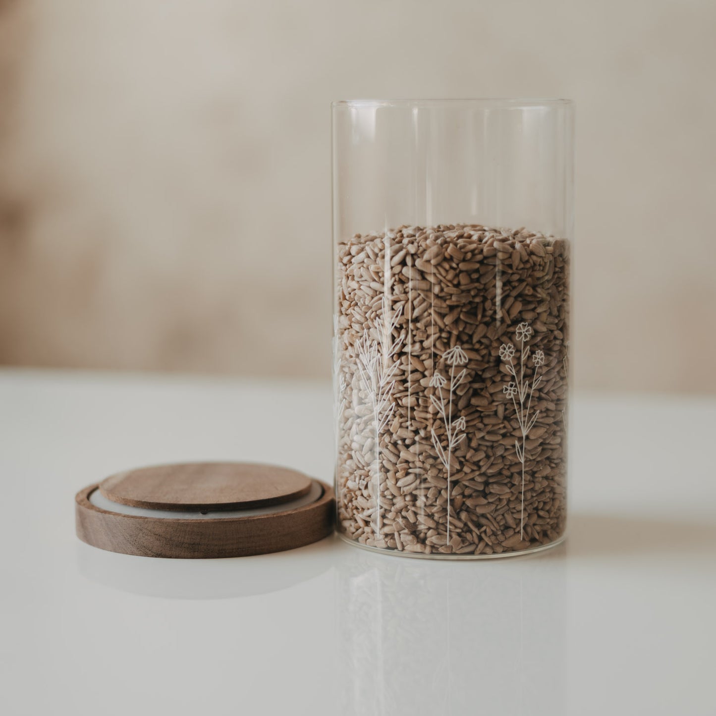 Geöffneter Glas-Vorratsbehälter mit weißem Wildblumen-Motiv, gefüllt mit Sonnenblumenkernen, daneben der Holzdeckel