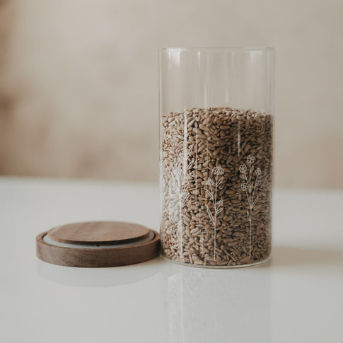 Geöffneter Glas-Vorratsbehälter mit weißem Wildblumen-Motiv, gefüllt mit Sonnenblumenkernen, daneben der Holzdeckel