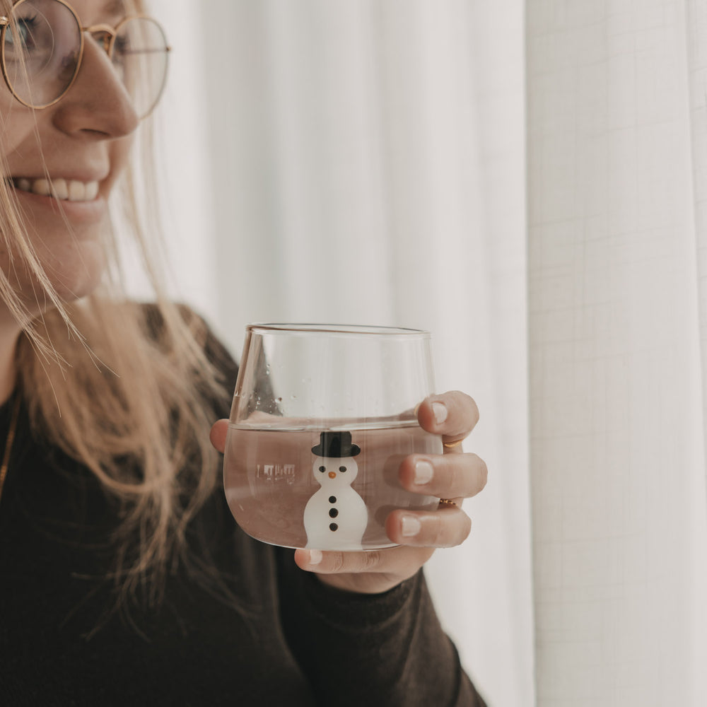 Frau mit Brille hält ein mit Wasser gefülltes Schneemann-Glas mit dekorativer Schneemannfigur im Glasboden.