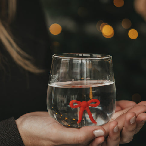 Transparentes Wasserglas mit roter Schleife im Glasboden, gehalten vor stimmungsvollen Weihnachtslichtern.