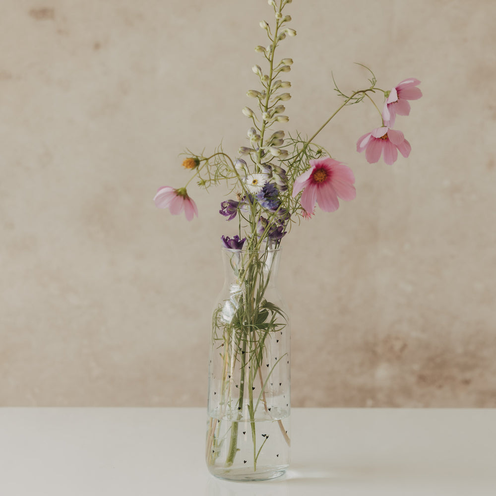 Glaskaraffe mit kleinen schwarzen Herzen als Vase mit rosa und violetten Blumen
