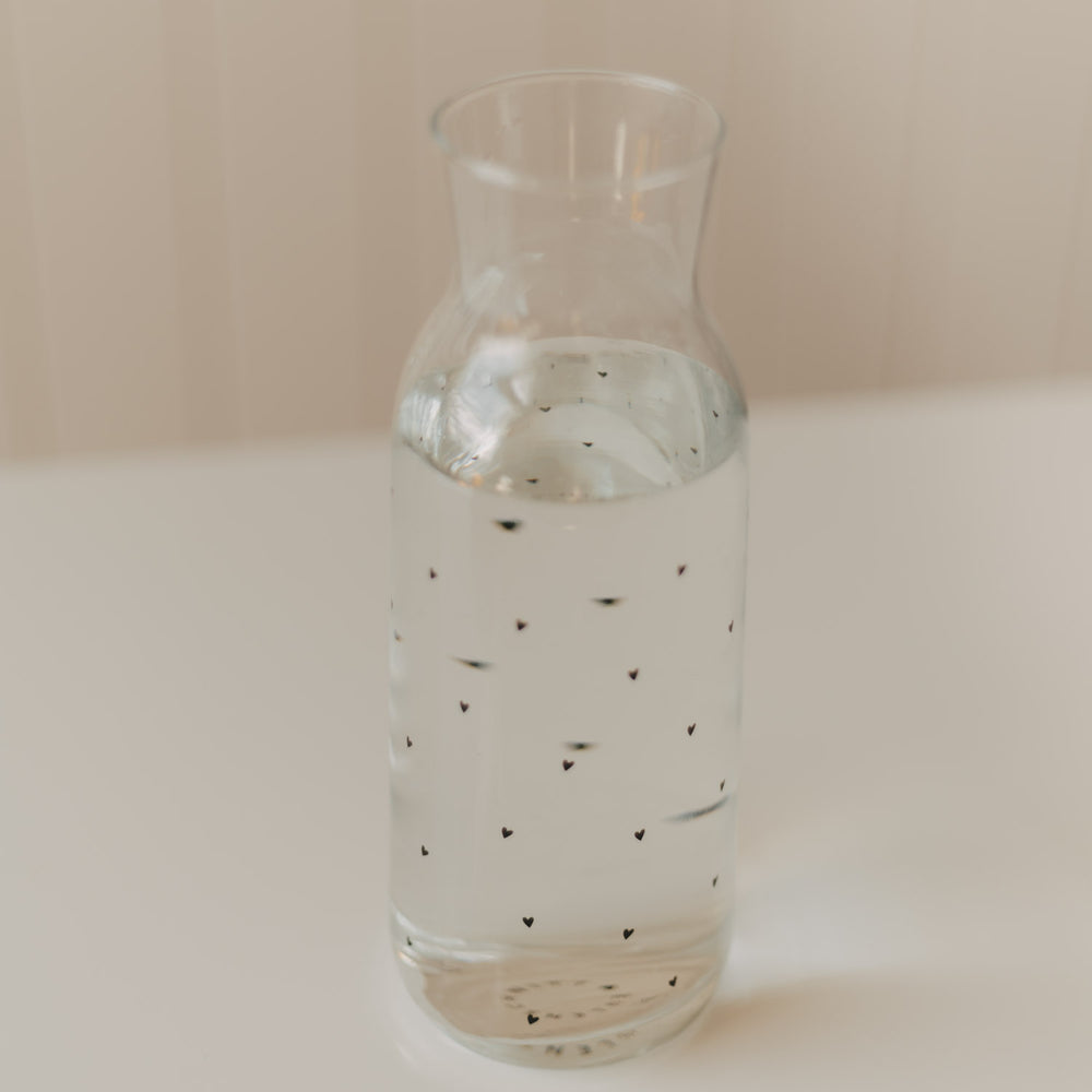 Mit Wasser gefüllte Glaskaraffe mit kleinen schwarzen Herzen auf hellem Untergrund