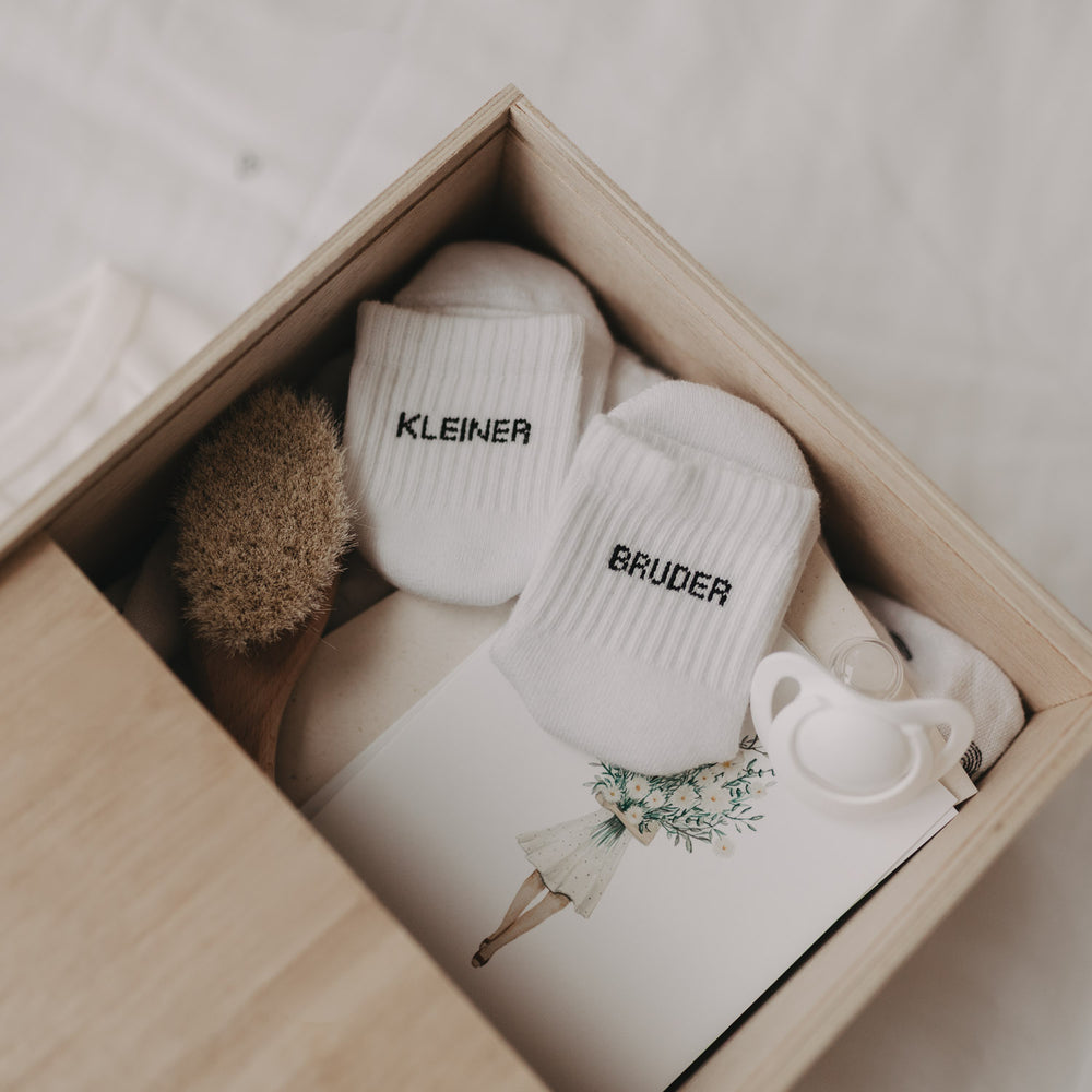 Offene Erinnerungsbox mit Baby-Erinnerungsstücken wie Söckchen „Kleiner Bruder“, Bürste und Schnuller.