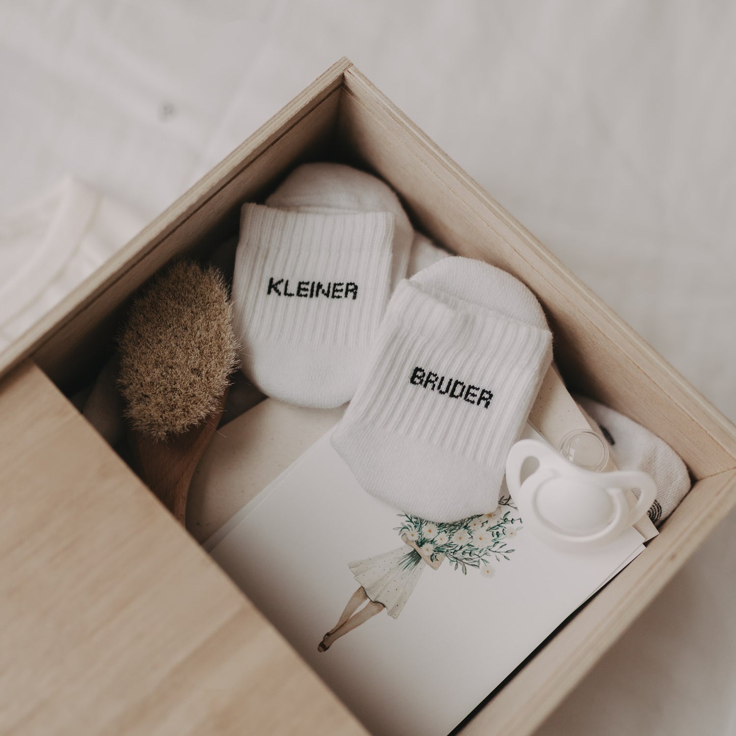 Offene Erinnerungsbox mit Baby-Erinnerungsstücken wie Söckchen „Kleiner Bruder“, Bürste und Schnuller.
