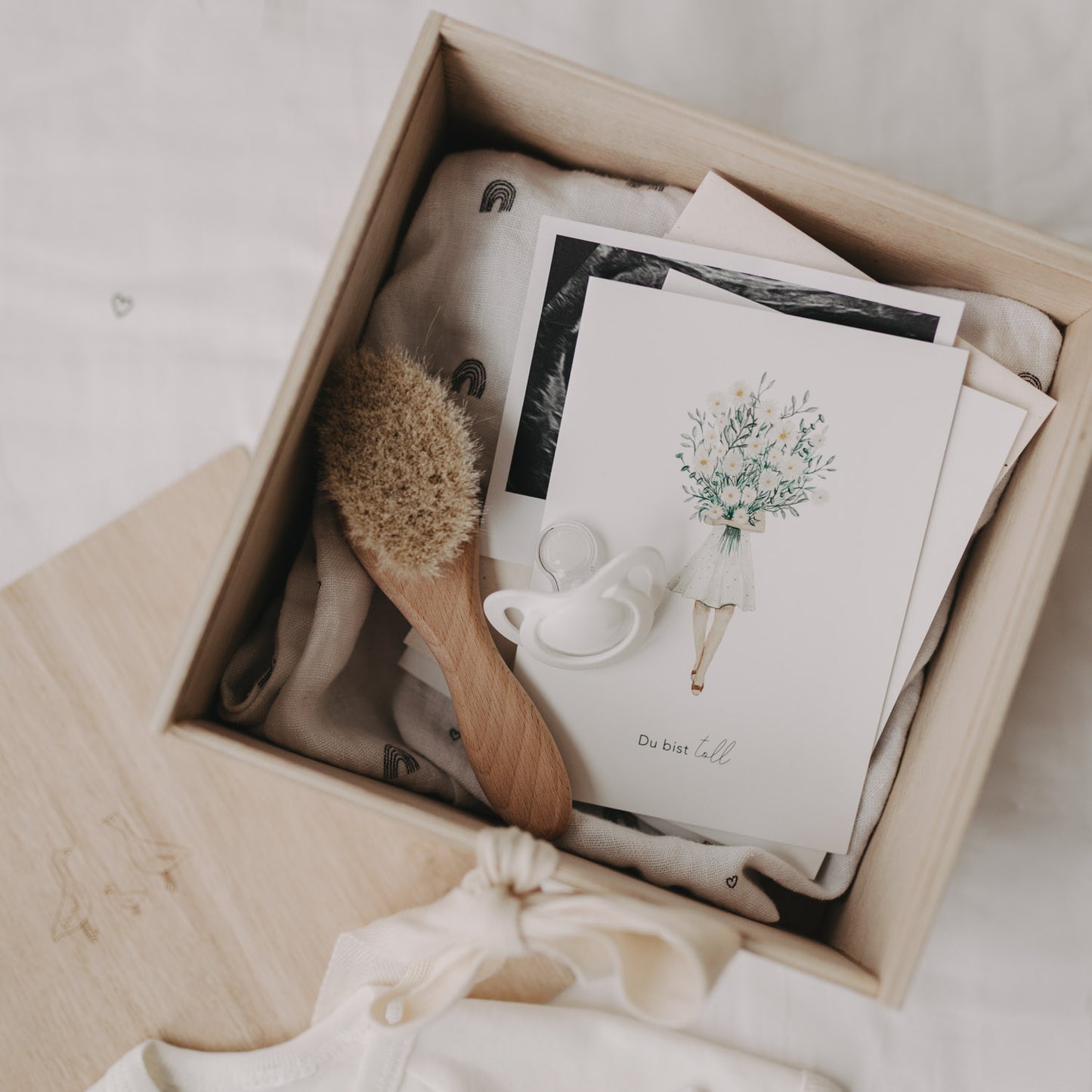 Geöffnete Erinnerungsbox gefüllt mit Babyartikeln wie Karte, Schnuller, Bürste und Tuch mit Regenbogenmotiv.