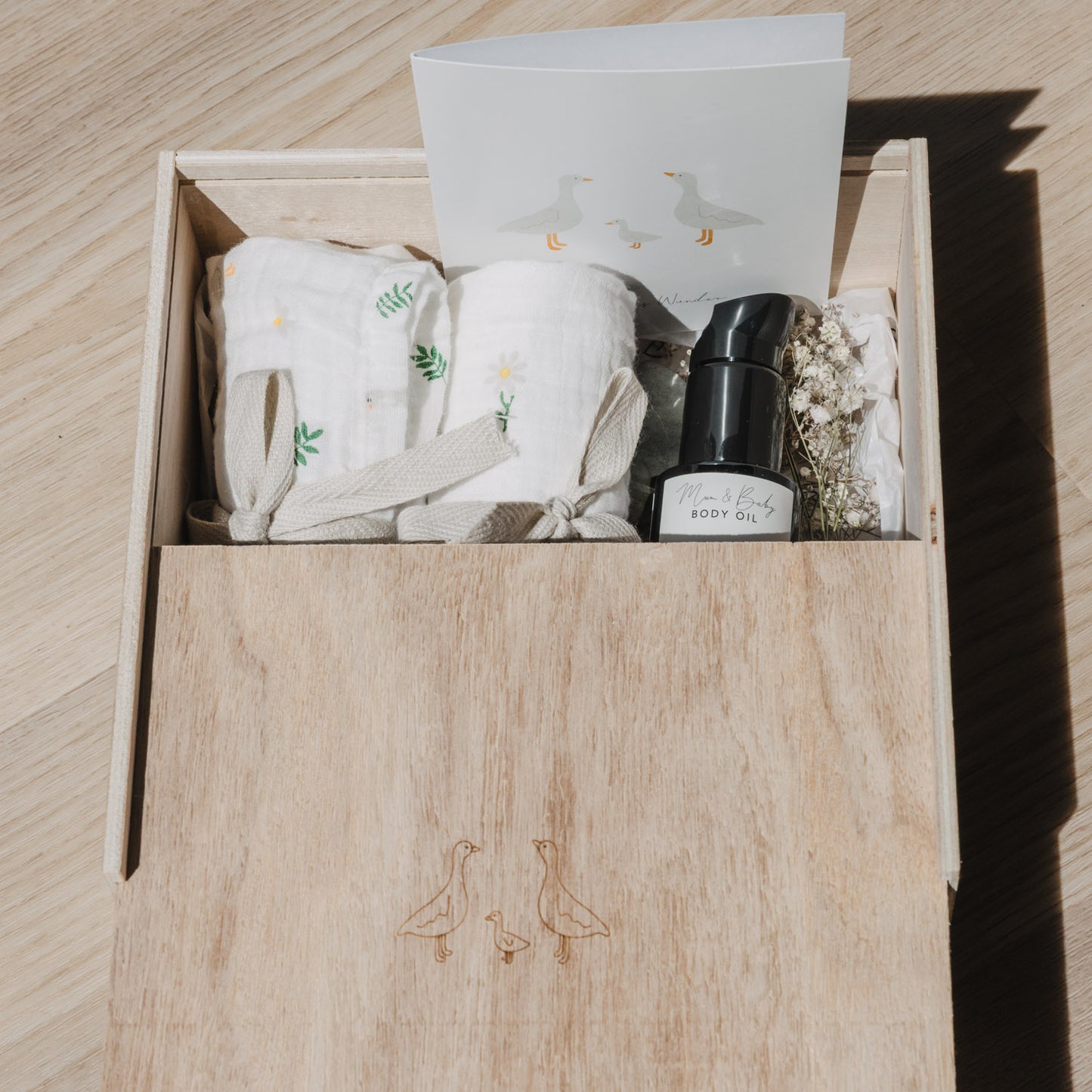 Geöffnete Erinnerungsbox aus Holz mit gravierter Gänsefamilie auf dem Schiebedeckel. Die Box ist mit Musselintüchern, einer Grußkarte, Trockenblumen und einem Pflegeöl gefüllt und eignet sich ideal als Geschenk zur Geburt oder Babyparty.