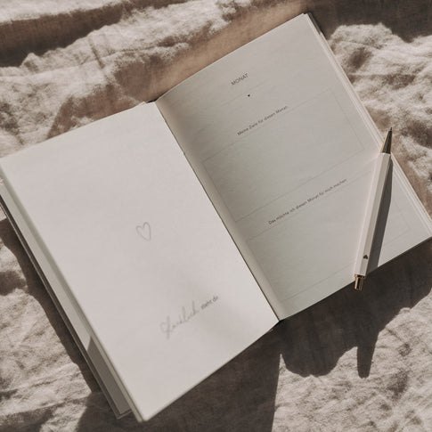 Offene Monatsseite des Kalenders Neues Glück mit Feldern für Ziele und Notizen, daneben ein Stift.