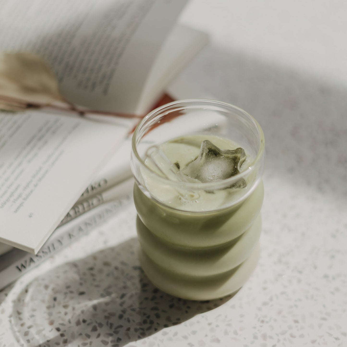Eulenschnitt Bubble Glas mit Iced Matcha Latte, darin ein sternförmiger Eiswürfel. Im Hintergrund liegt ein geöffnetes Buch auf einer hellen Oberfläche.