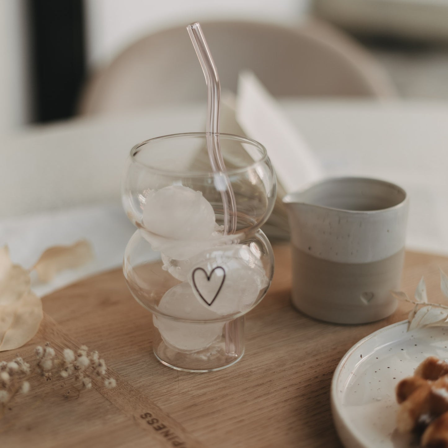 Eulenschnitt Bubble Glas mit schwarzem Herz-Print, gefüllt mit Iced Coffee und Eiswürfeln. Daneben ein Keramikkännchen und ein Teller mit belgischer Waffel auf einem runden Holzbrett.