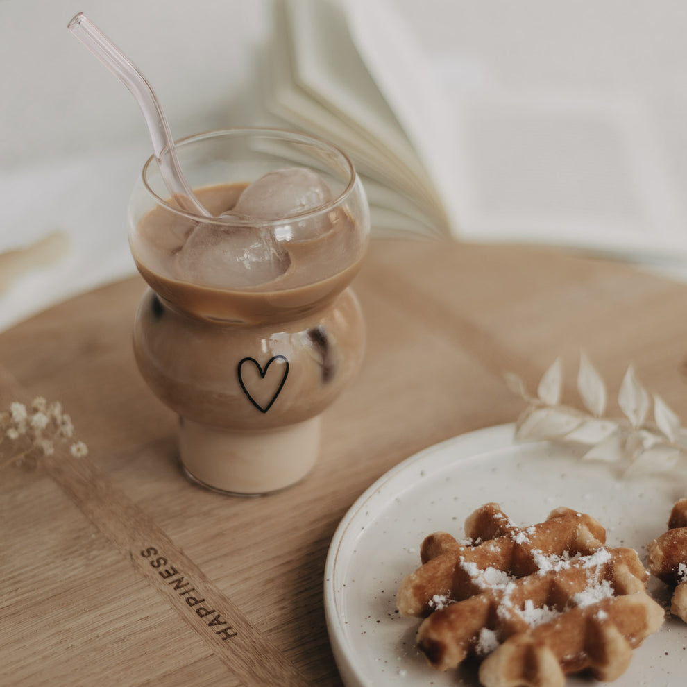 Eulenschnitt Bubble Glas mit schwarzem Herz-Print, gefüllt mit Iced Coffee und runden Eiswürfeln. Daneben ein kleiner Keramikteller mit belgischer Waffel auf einem runden Holzbrett