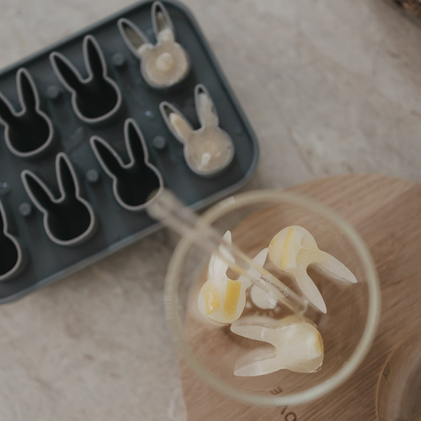 Ansicht von oben der grauen Eiswürfelform aus Silikon mit erhöhten Hasenformen, 2 davon gefüllt mit Eis. Ein Glas mit Hasen-Eiswürfeln und Strohhalm steht daneben.
