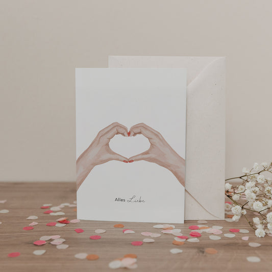 Klappkarte mit zwei Händen in Herzform und dem Schriftzug „Alles Liebe“, vor Umschlag und Blumen, auf hellem Untergrund.