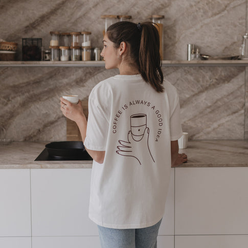 Frau steht in Küche, hält Kaffeetasse und trägt T-Shirt mit Illustration und Text „Coffee is always a good idea“.