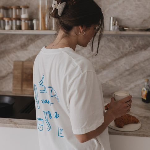 Frau mit Frühstücksshirt bereitet Kaffee zu, Rückseite zeigt blaue Frühstücksmotive.