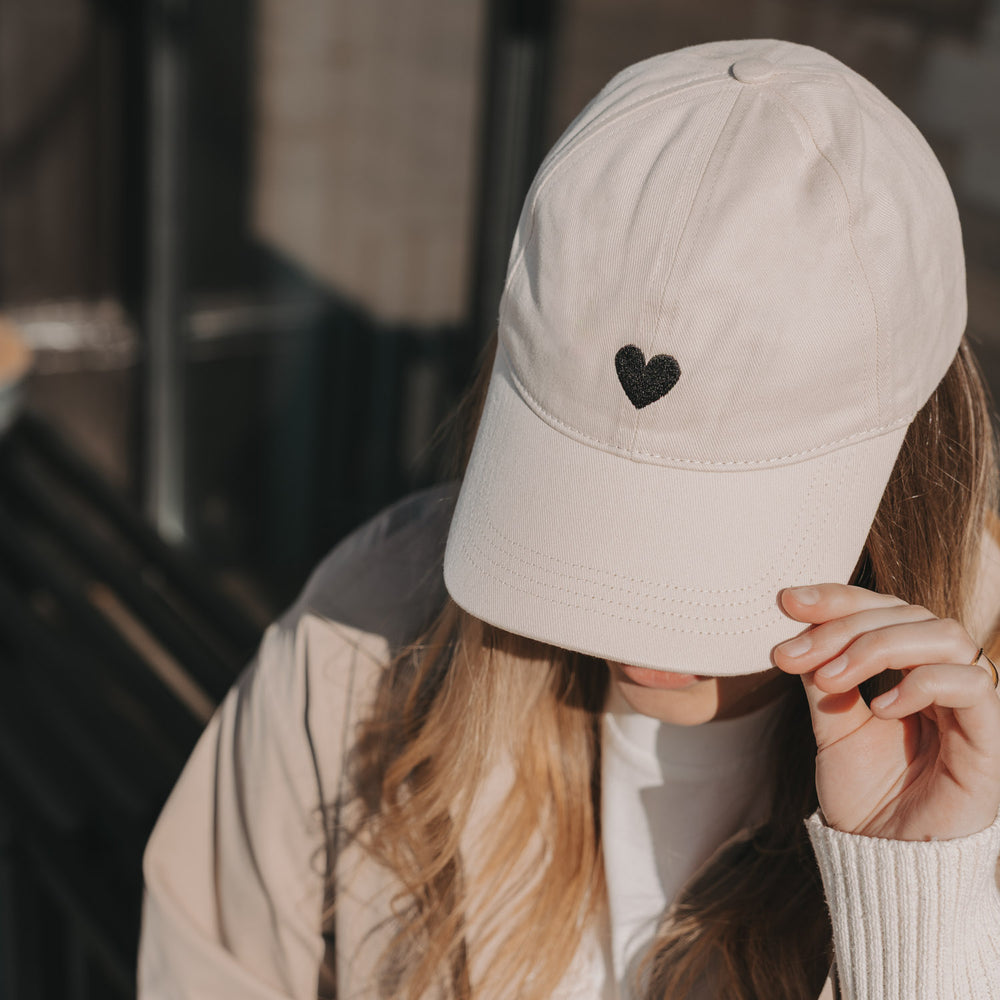 Beige Baseball Cap mit schwarzem Herz-Stick in Händen gehalten