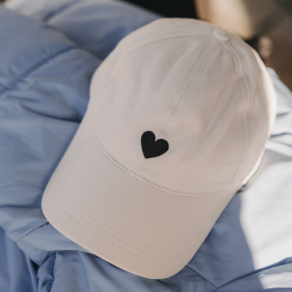 Beige Cap mit schwarzem Herz-Stick liegend auf blauer Jacke