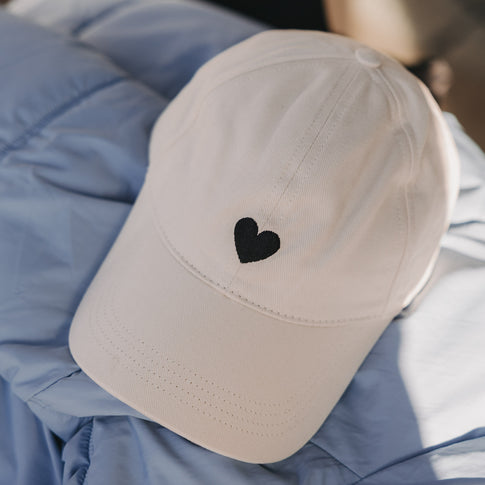Beige Cap mit schwarzem Herz-Stick liegend auf blauer Jacke
