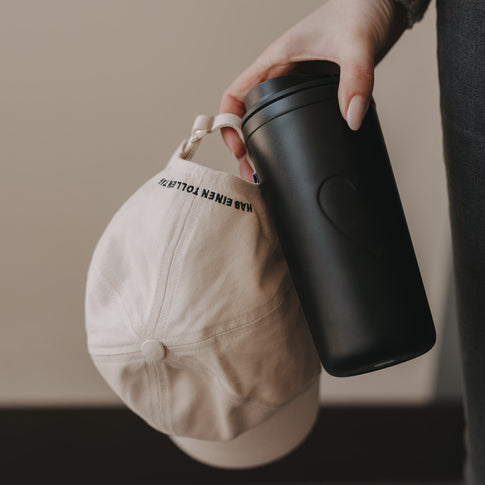 Hand hält beige Cap mit Herz-Stick neben schwarzem Thermobecher